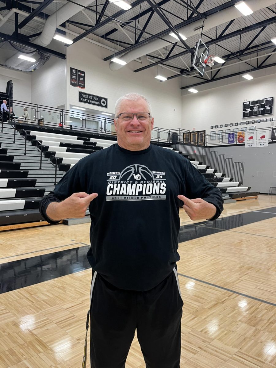 Coach Paul Chapman smiling proud in his home gymnasium pointing to his district and regional champion shirt.